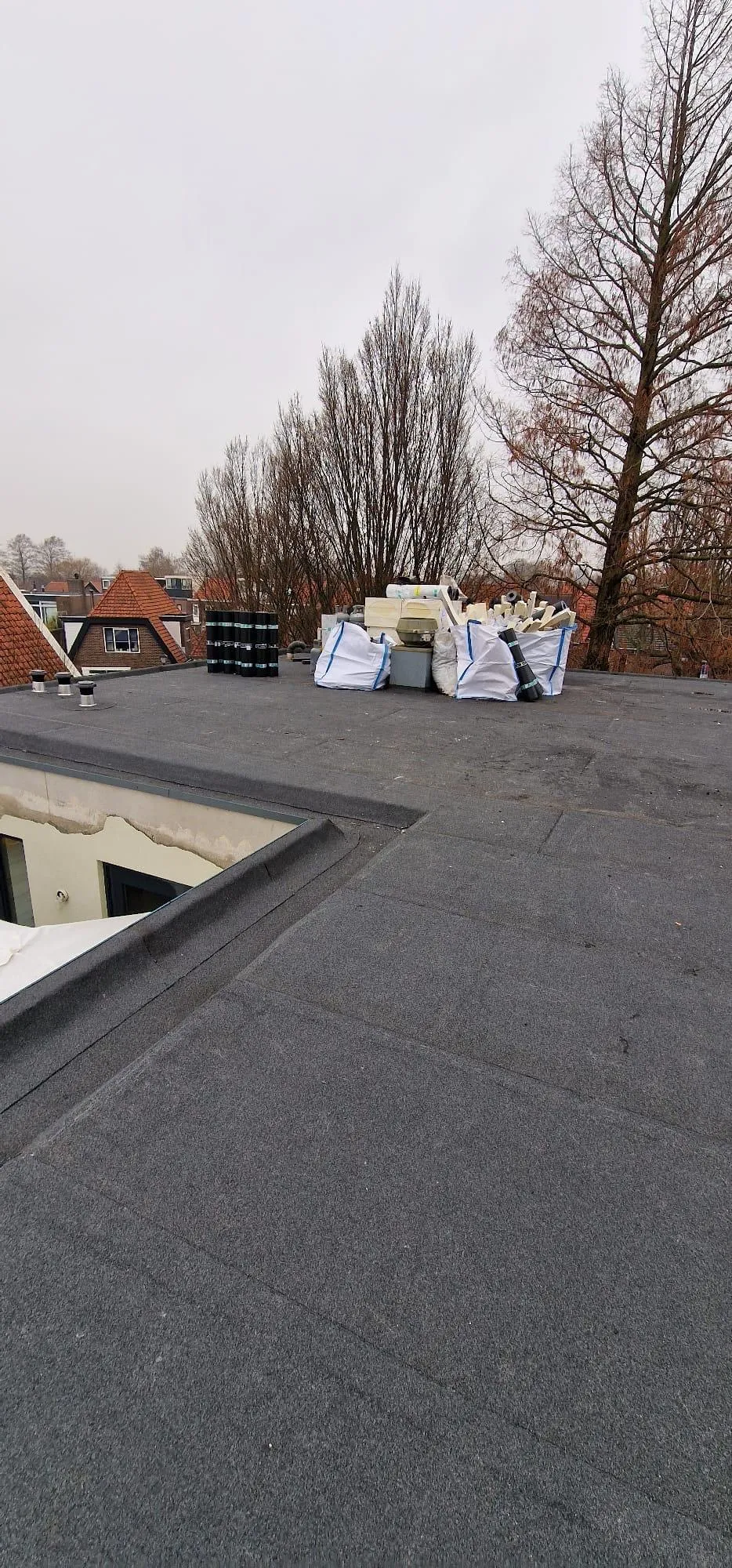 Plat dak met stapel bouwmaterialen zoals rollen dakbedekking en witte zakken bij kale bomen.