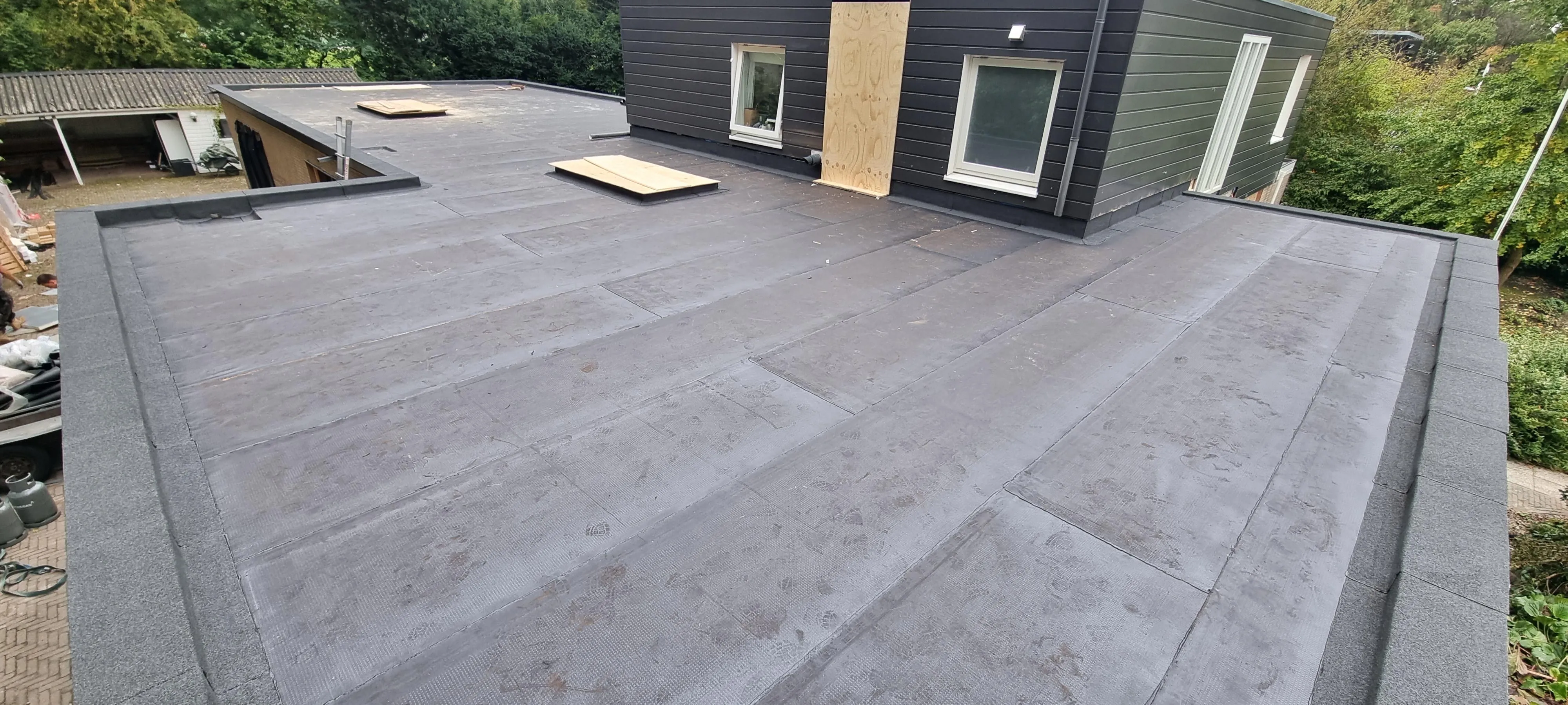 Flat roof under construction with waterproof membrane and two covered skylight openings near black siding walls.