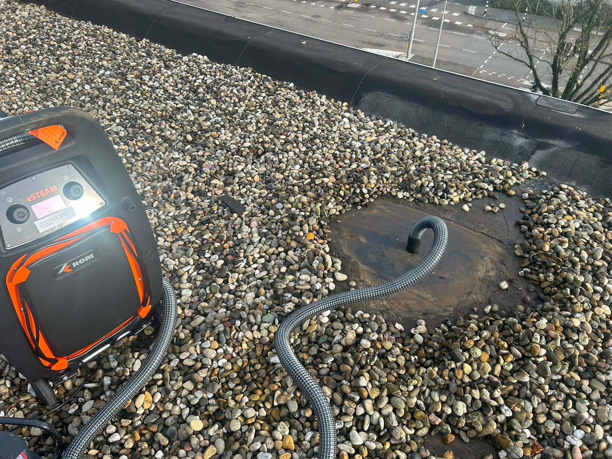 Black and orange eSTEAM device on a pebble-covered flat roof with a vent pipe and flexible hose attached.