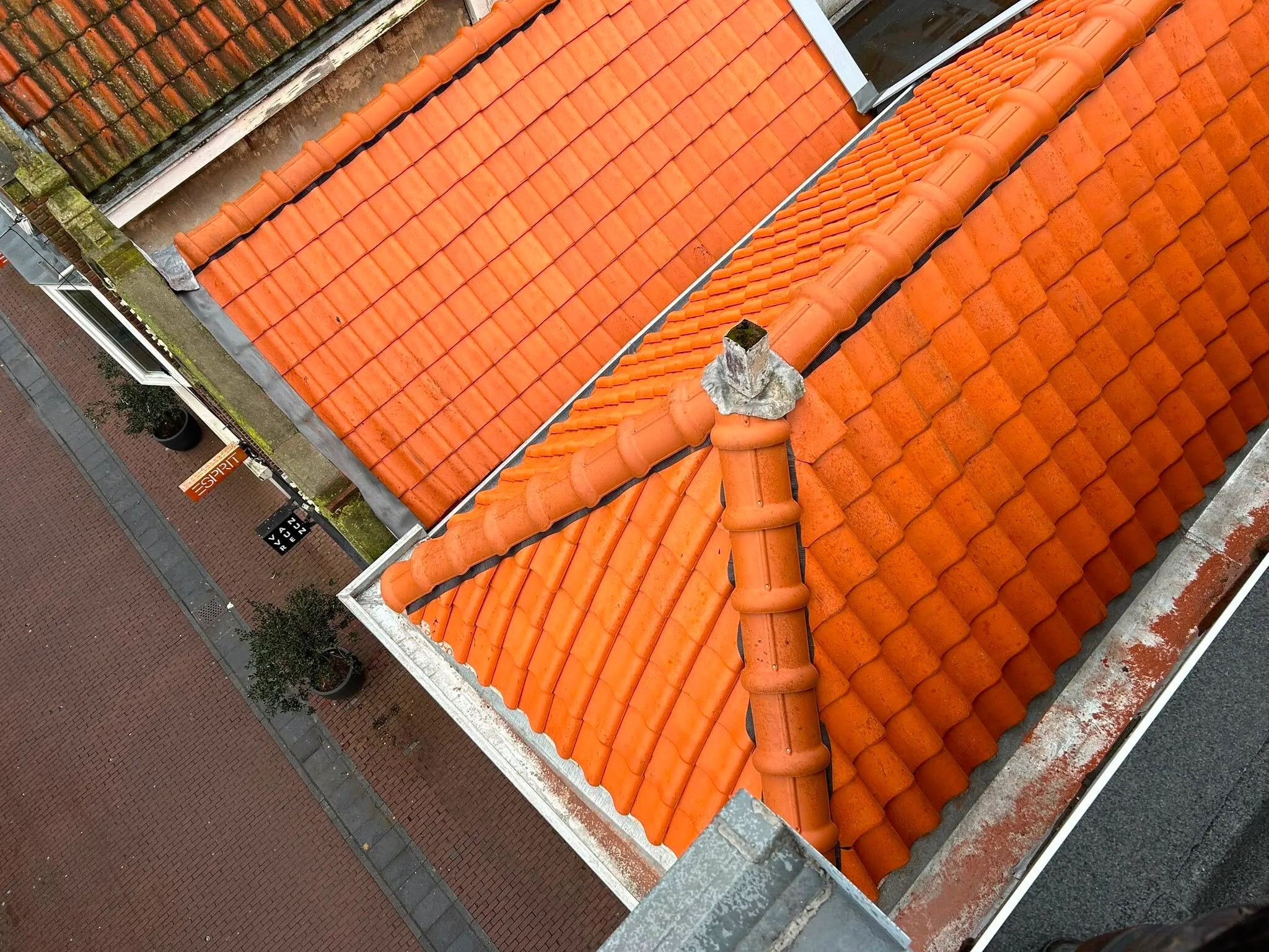 View of red-orange tiled rooftops next to a brick-paved street with potted plants and shop signs.