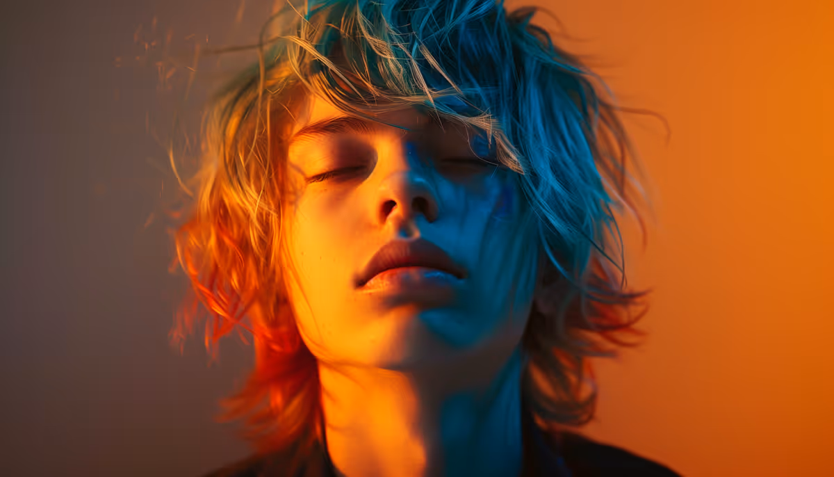 Close-up of a young person with closed eyes and tousled hair lit by warm orange and cool blue light.