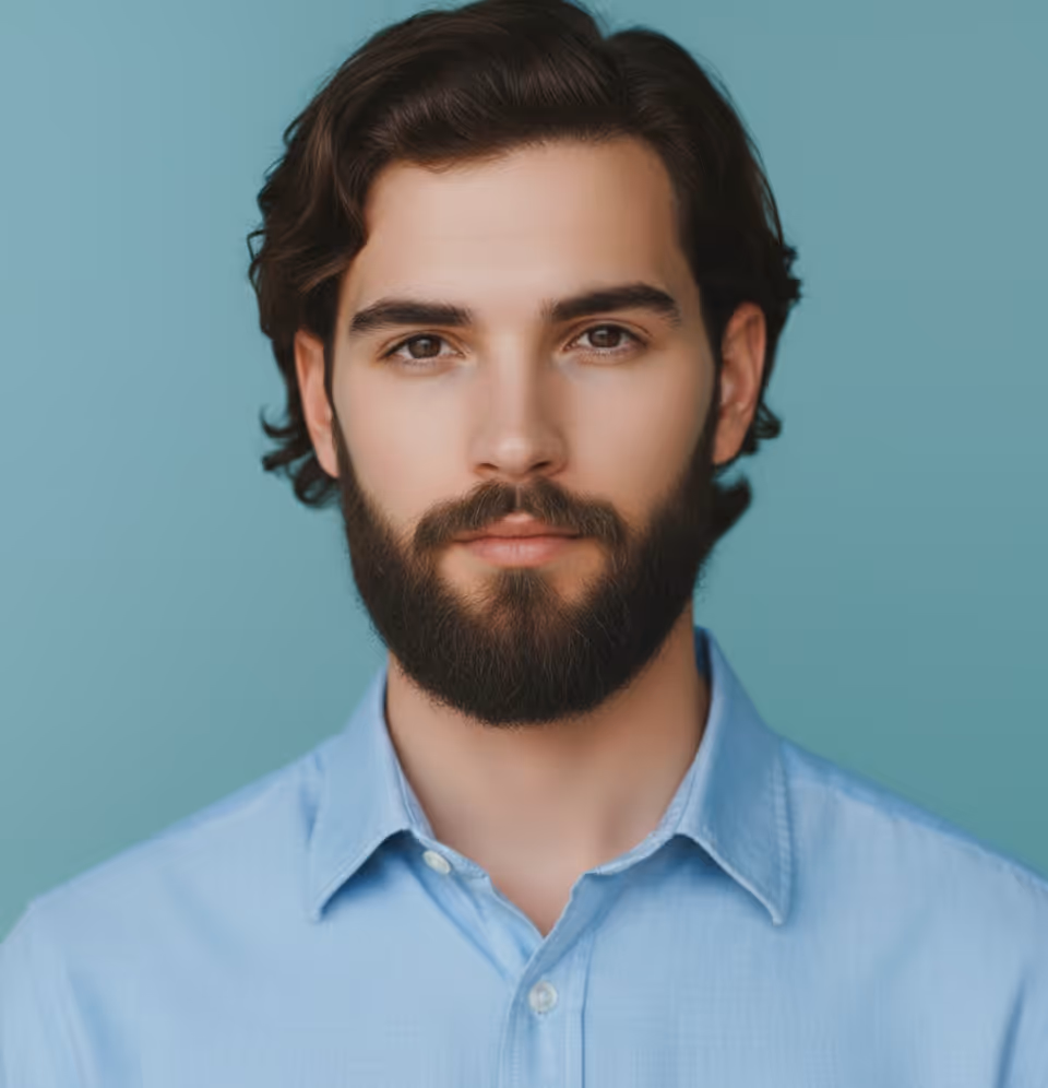 Young man with dark hair and beard wearing a light blue button-up shirt against a teal background.