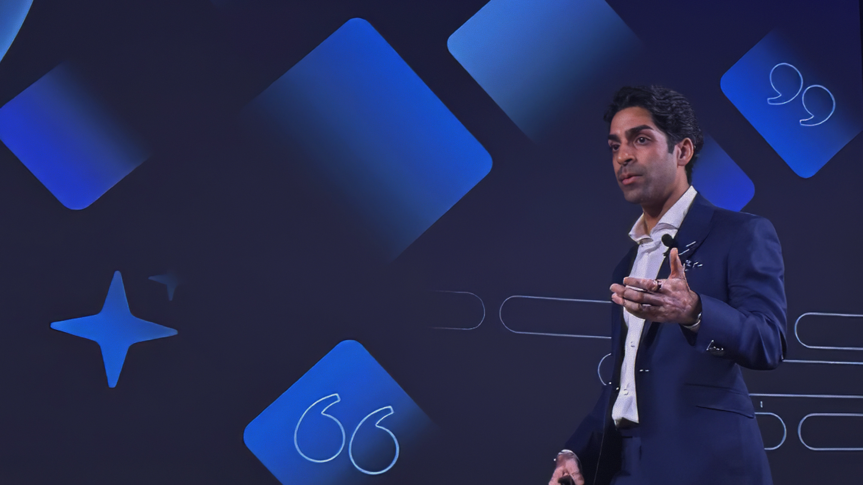 Man in a dark suit speaking on stage with blue geometric shapes and quotation marks in background.