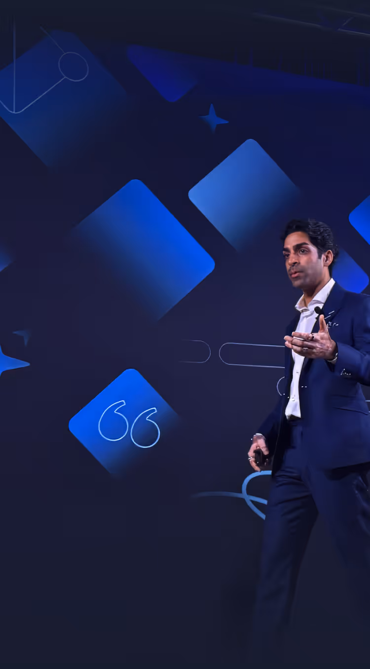 Man in blue suit speaking and gesturing on stage against a blue and black geometric background.