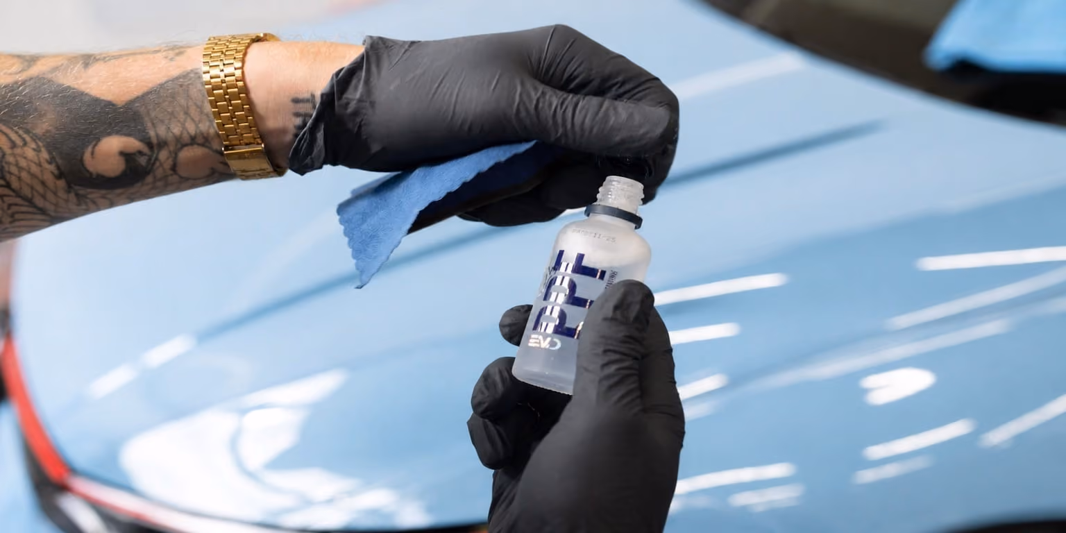 Person mit schwarzen Handschuhen hält eine geöffnete Flasche mit Flüssigkeit in der einen und ein blaues Tuch in der anderen Hand über einer hellblauen Autokarosserie.