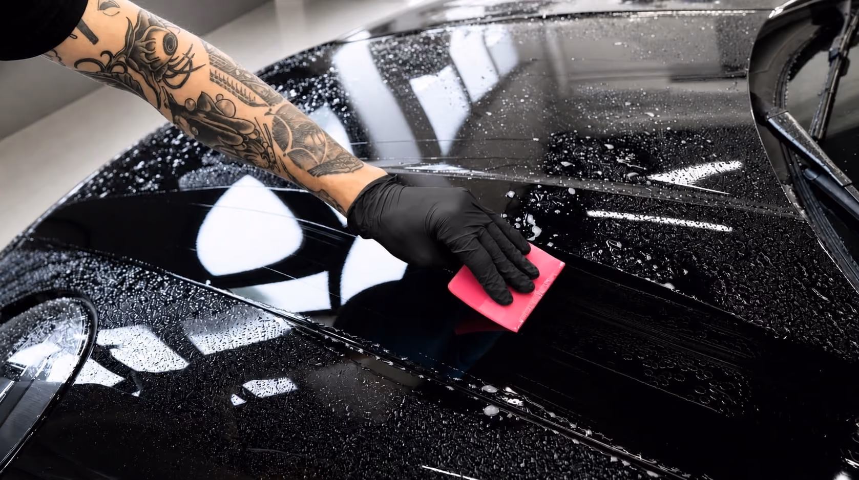 Eine tätowierte Hand mit schwarzem Handschuh reinigt die nasse Motorhaube eines schwarzen Autos mit einem rosa Schwamm.