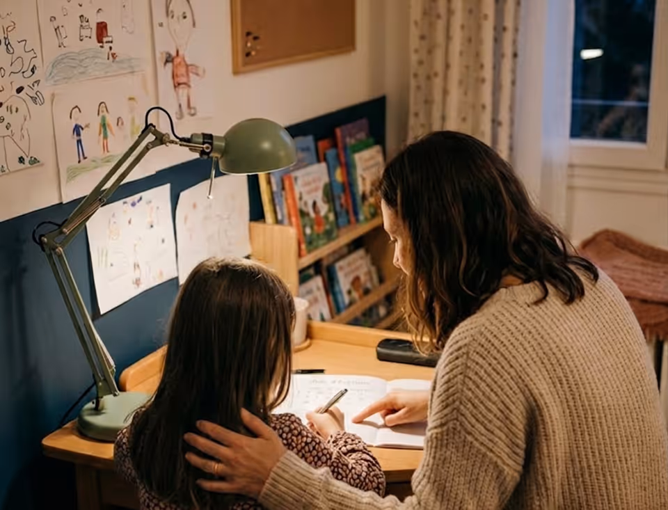 Une femme aide une fillette à faire ses devoirs à un bureau avec une lampe et des dessins accrochés au mur.