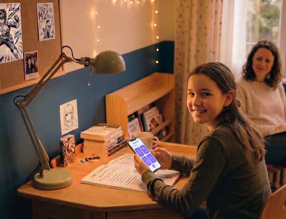 Une fille souriante avec un téléphone assise à un bureau, une femme regarde en arrière-plan.