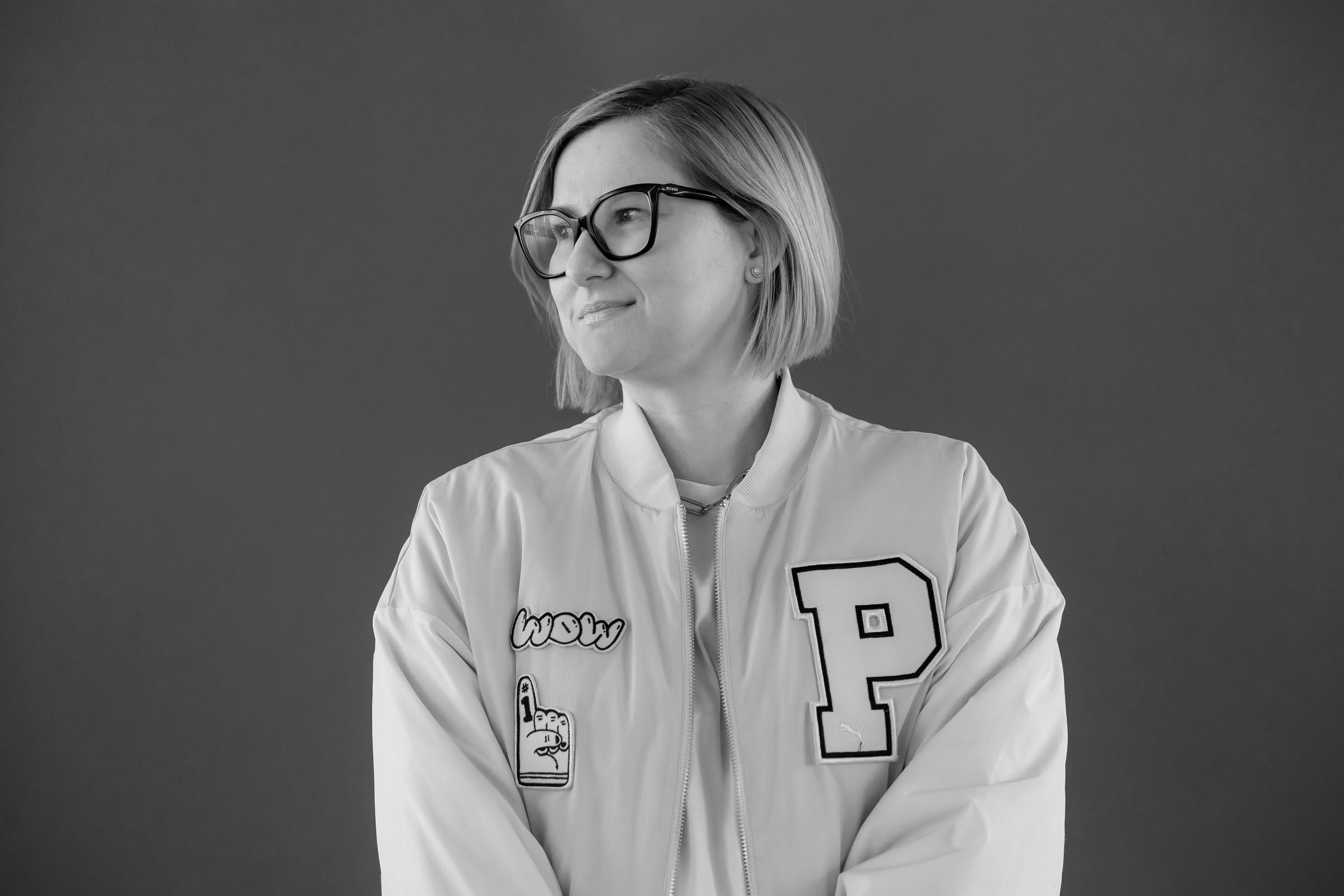 Woman with short hair and glasses wearing a jacket with patches, looking to her left.