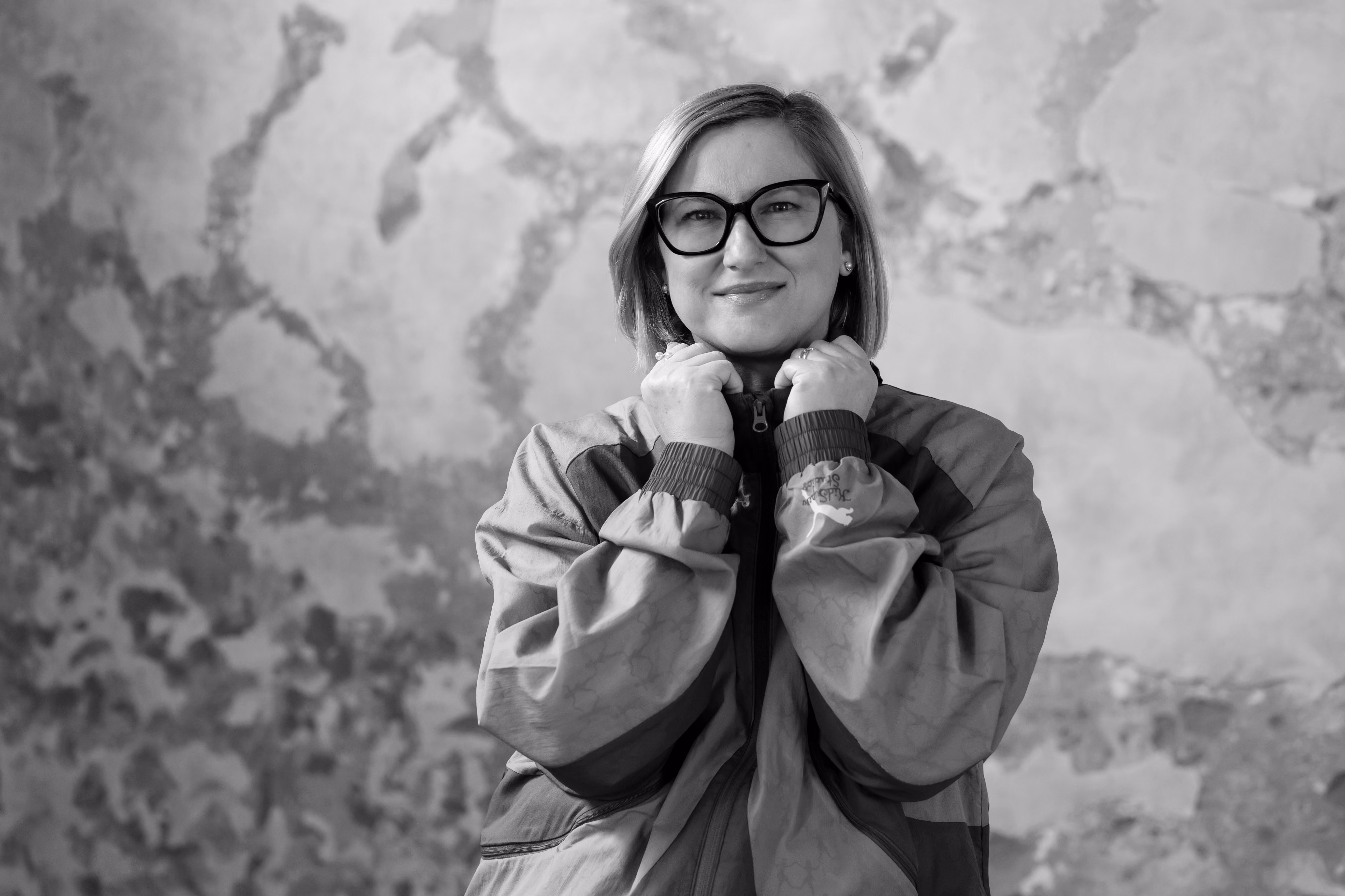 Smiling woman with short hair and glasses wearing a zippered jacket against a textured wall.