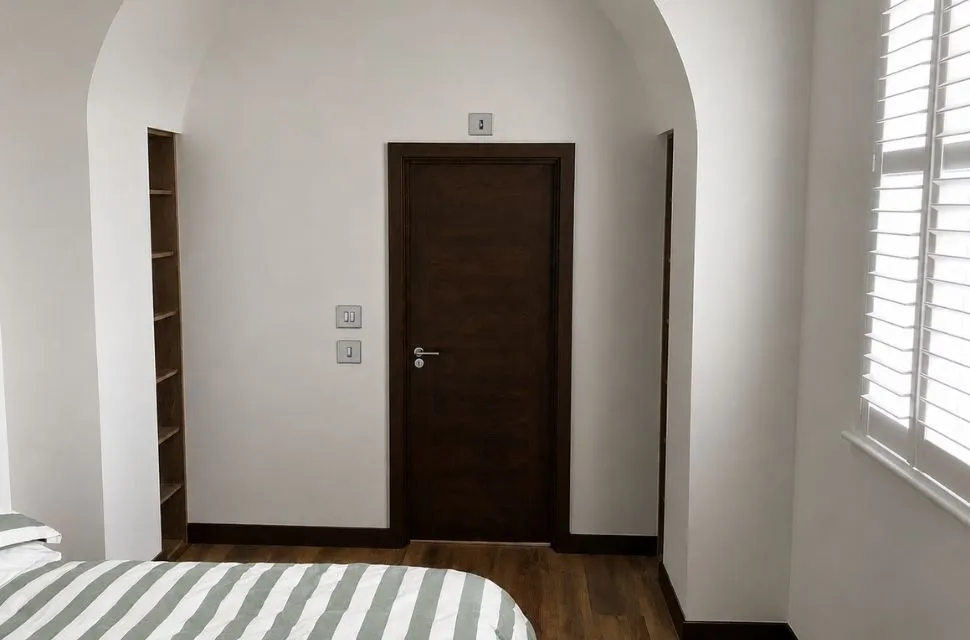 Minimalist bedroom with a striped bed, wooden door, built-in shelves, and white shuttered window.