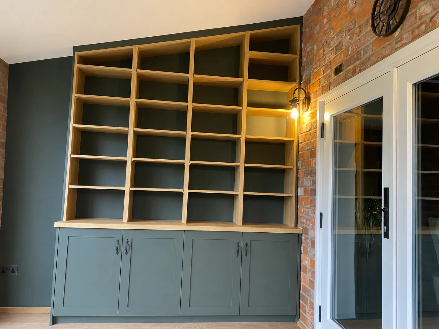 Empty wooden bookshelf with green lower cabinets against a dark wall and brick beside a glass door.