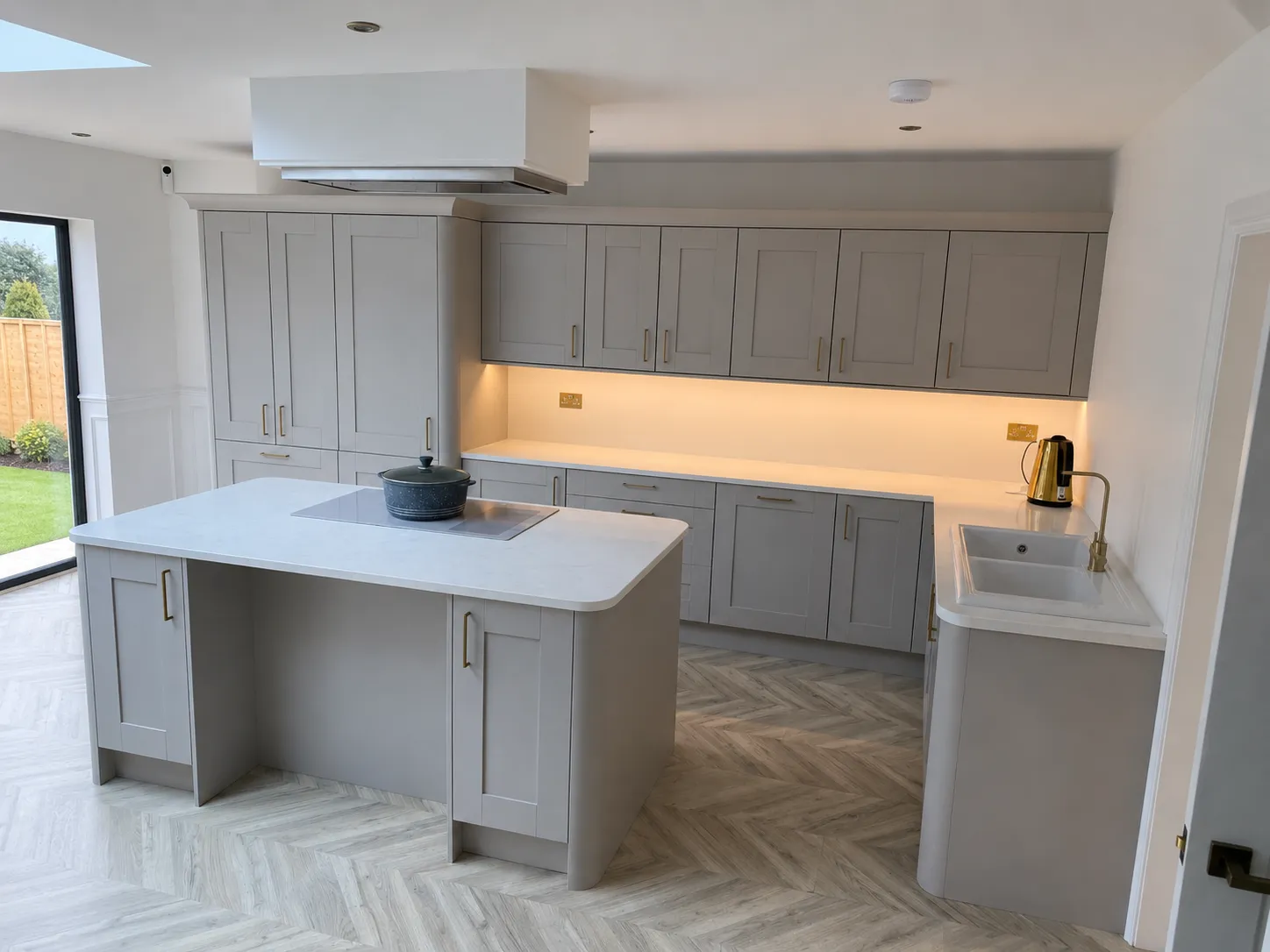 Modern kitchen with light gray cabinets, white countertops, island with stove, and a gold kettle near the sink.