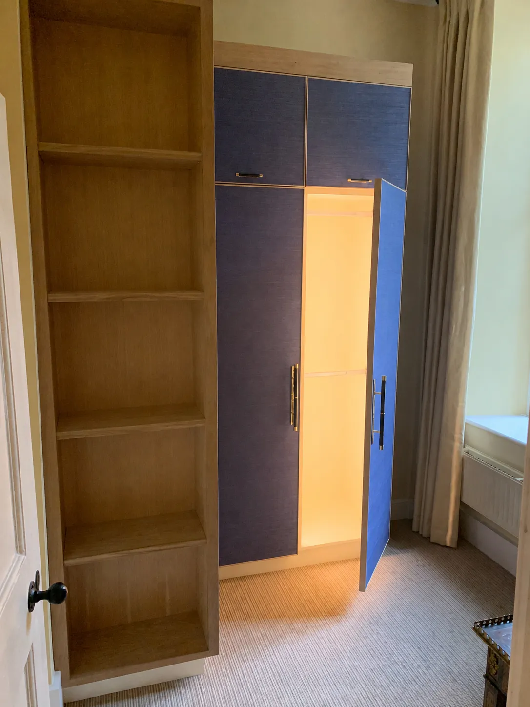 Room corner with open blue wardrobe door and adjacent empty wooden shelves, beige carpet, and curtain.