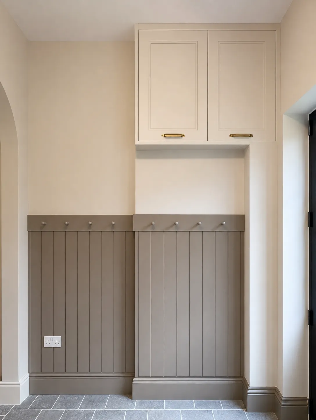 Minimalist hallway with beige walls, taupe paneling with hooks, white cabinet, and stone tile floor.