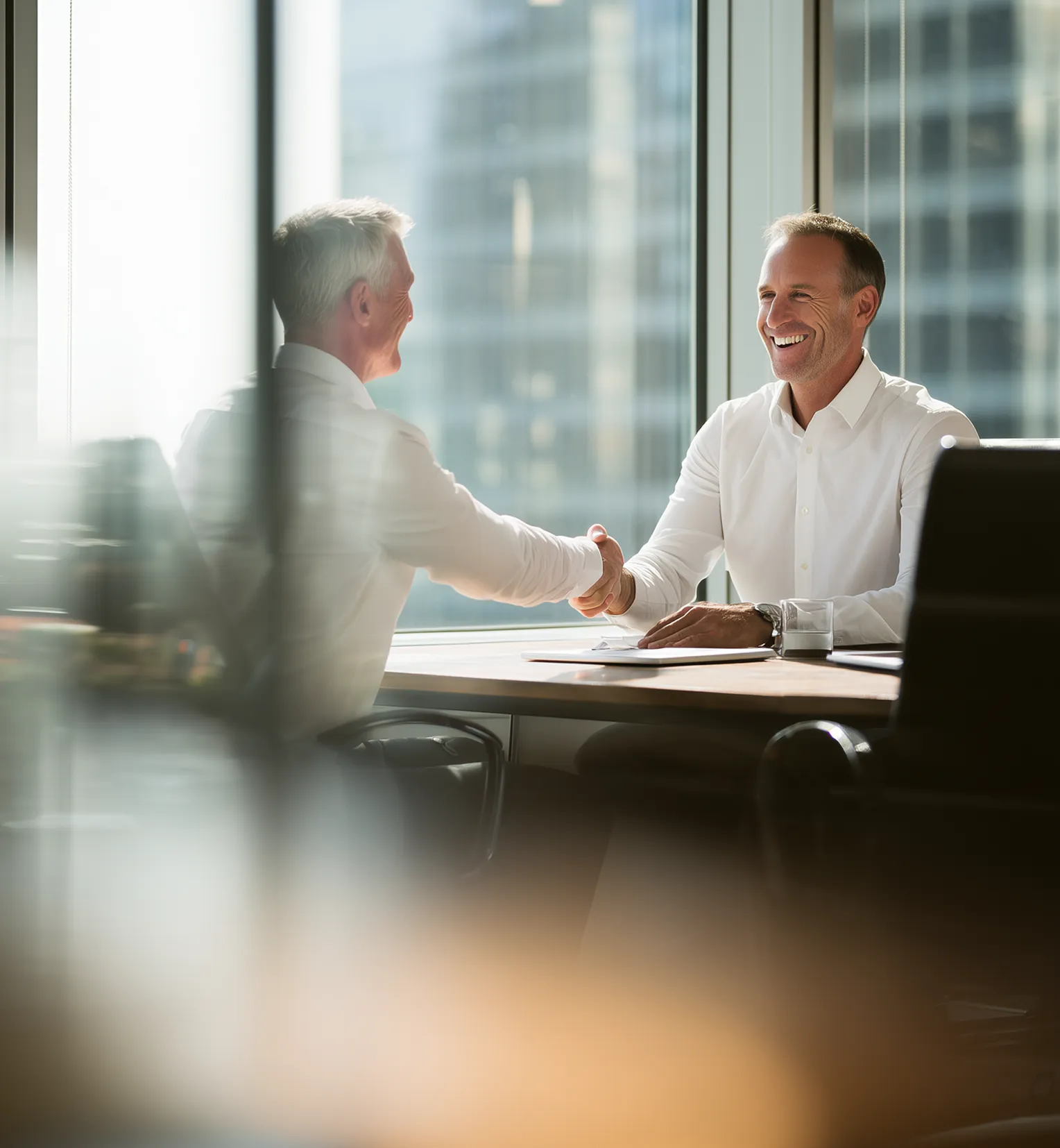 Two men in white shirts shaking hands and smiling in a bright office with large windows.