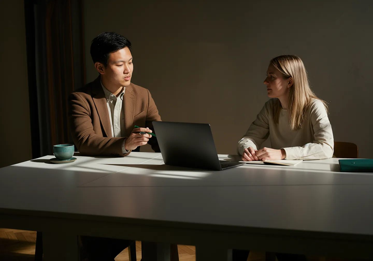 Man in brown jacket and woman discuss over laptop at white table with notebook and cup.