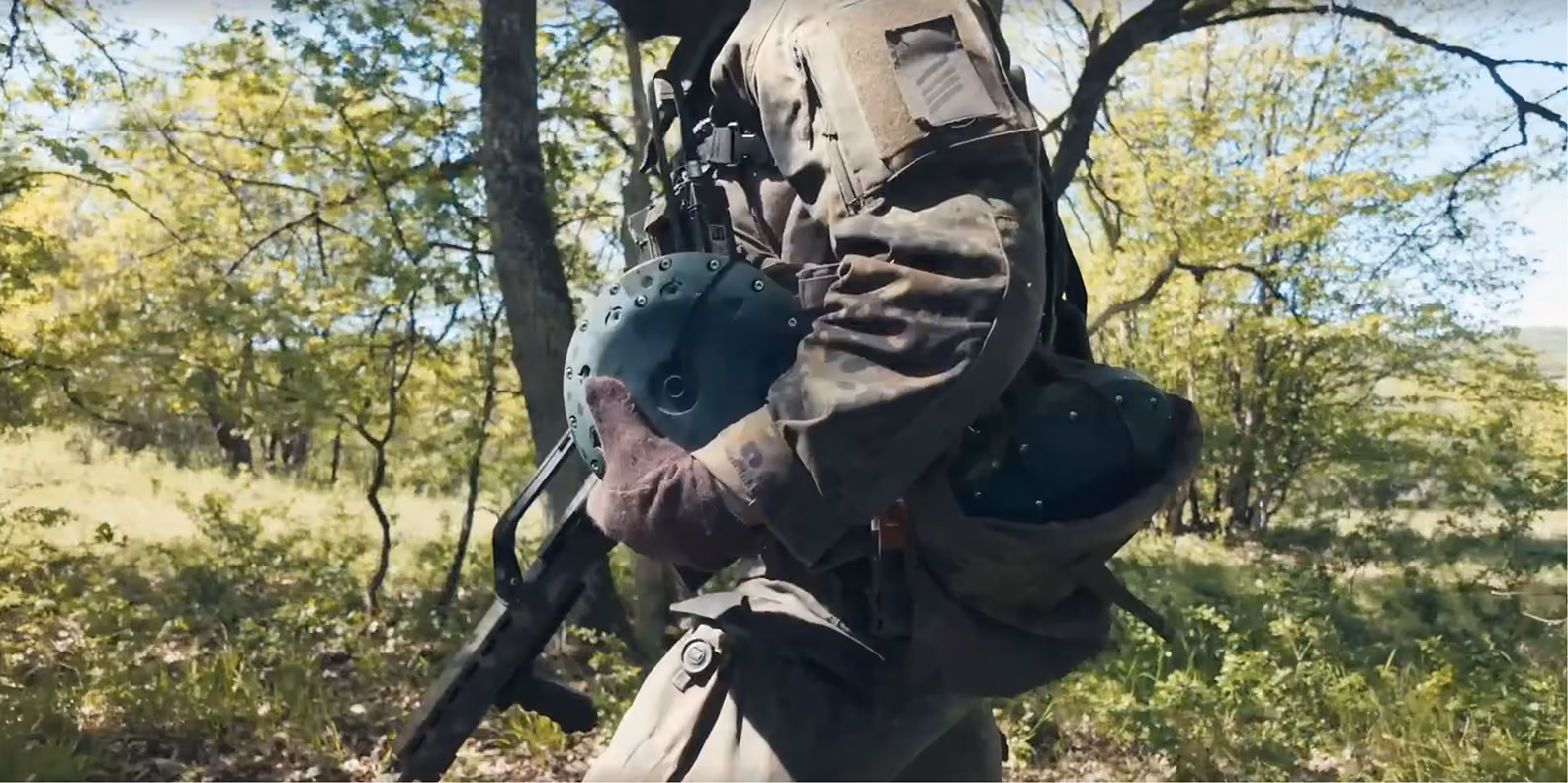 Soldier in camouflage holding a helmet and rifle walking through a sunlit forest area.