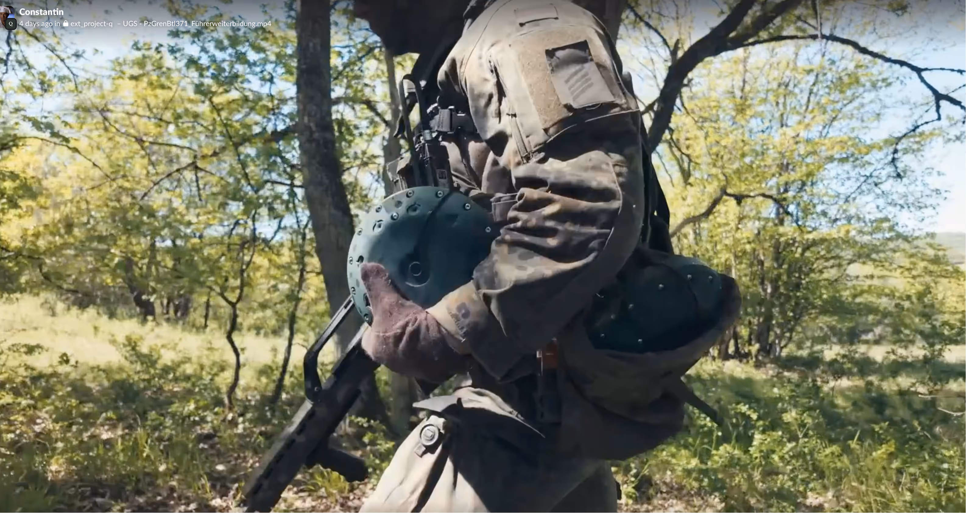 Soldier in camouflage holding a helmet and rifle walking through a sunlit forest area.
