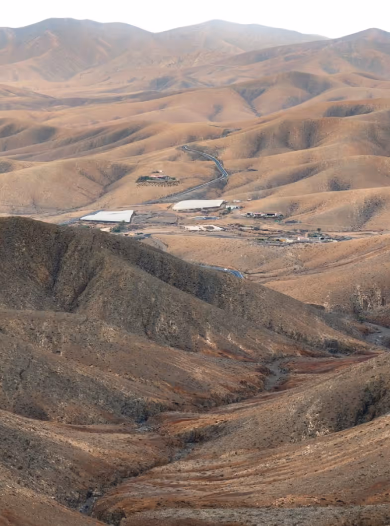 Arid, hilly landscape with scattered buildings and winding roads under bright sky.