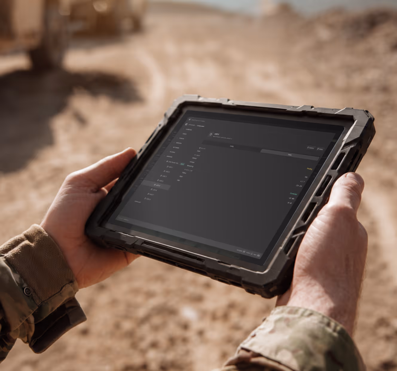 Person in camouflage holds rugged tablet displaying a dark-themed interface in a sandy outdoor setting.