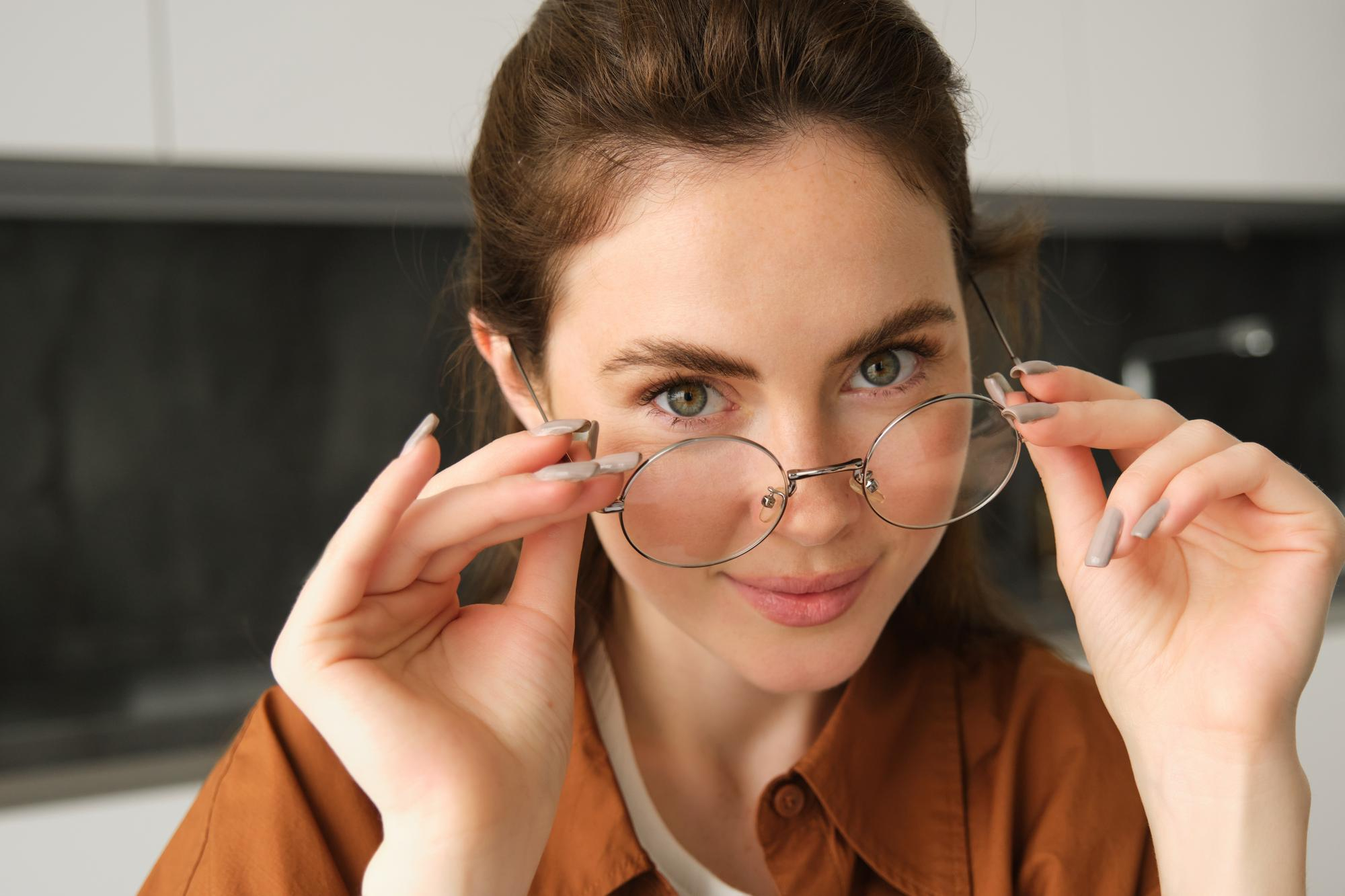 Woman with green eyes wearing round glasses and a brown shirt, holding the glasses with both hands.