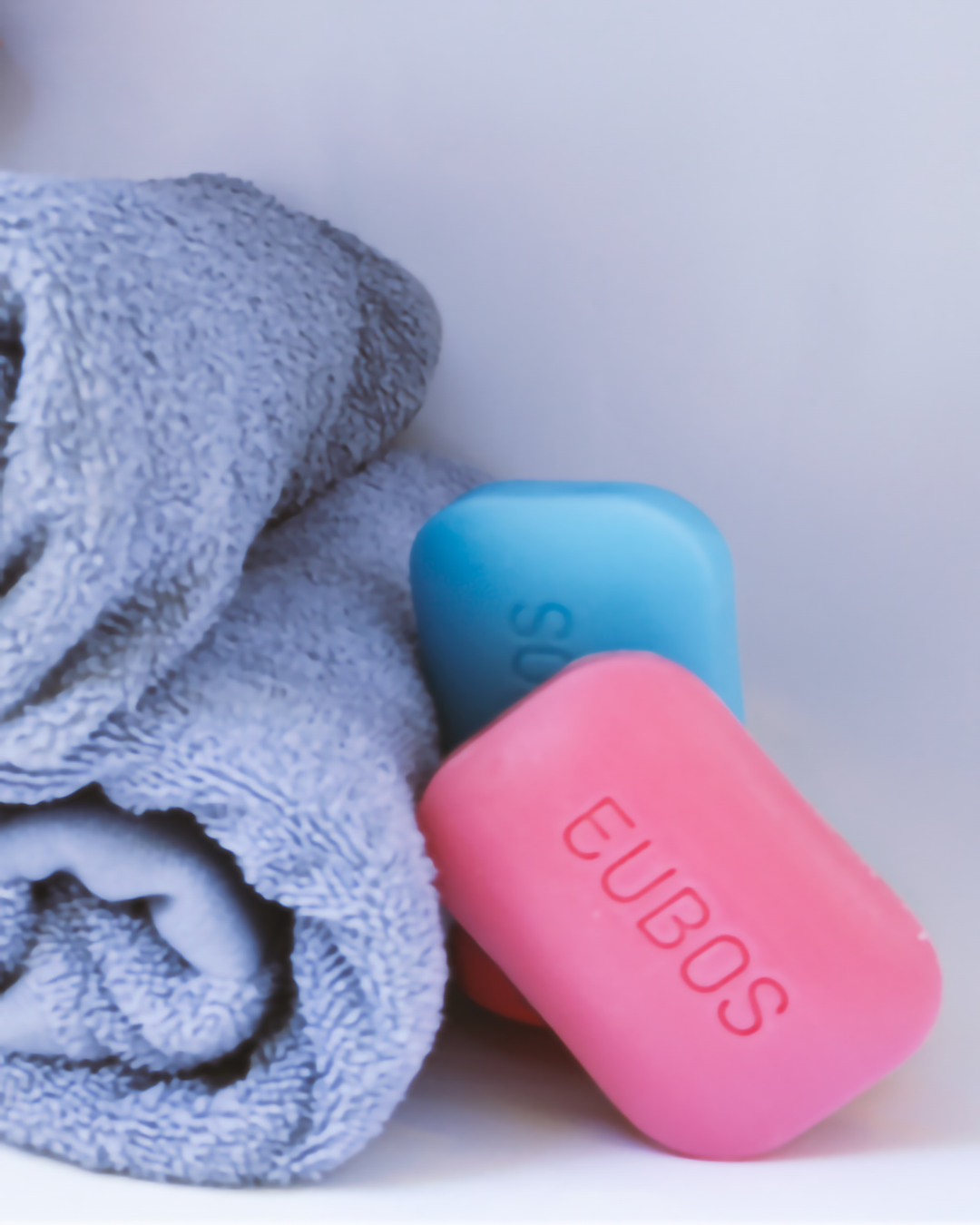 Pink and blue Eubos soap bars beside a rolled towel in soft pastel lighting.