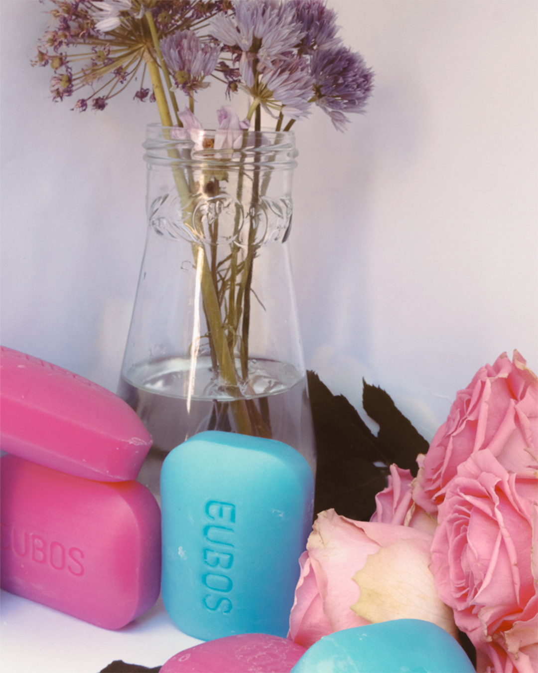 Pink and blue Eubos soap bars surrounded by flowers and a glass vase.