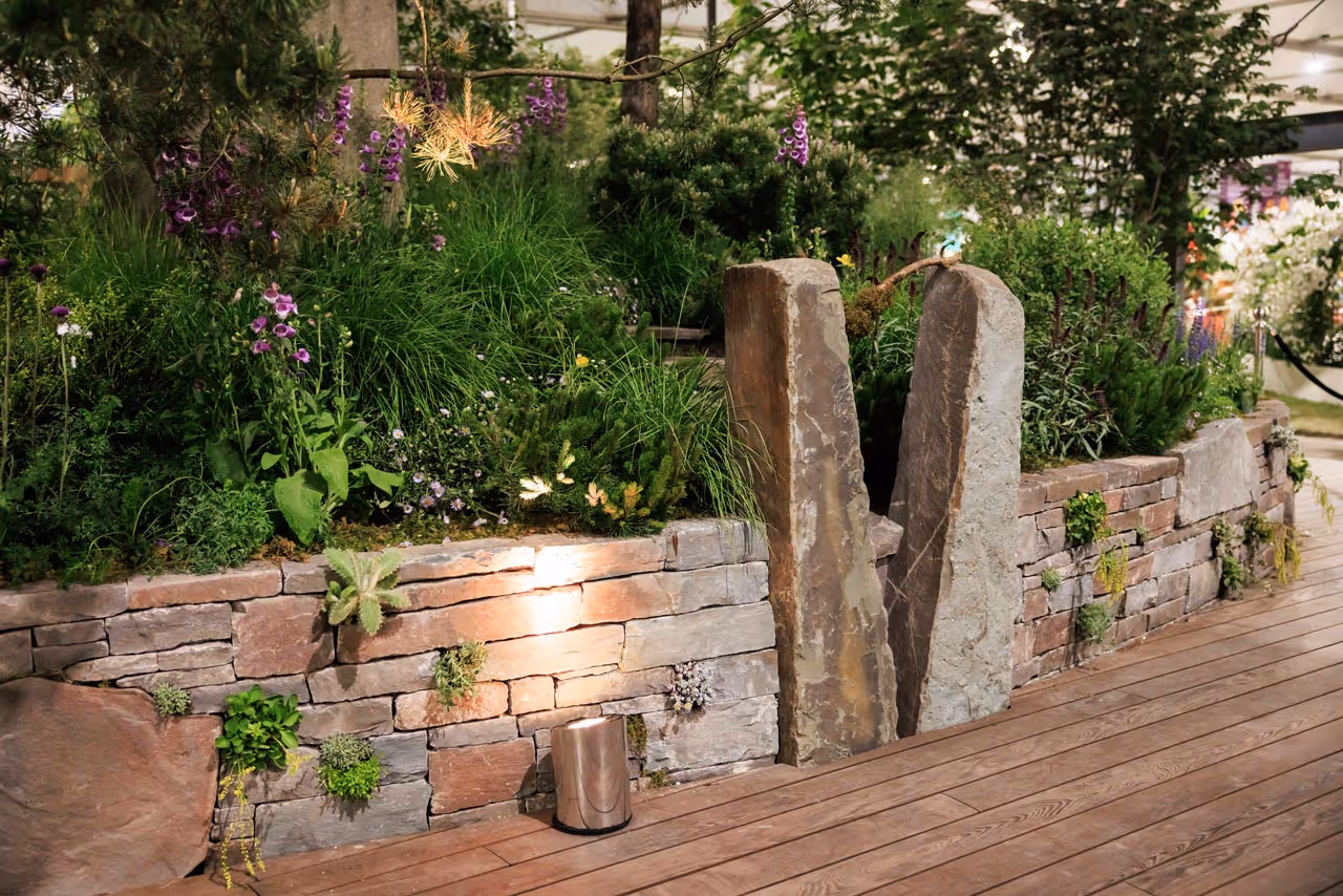 Stone planter with shrubs and purple flowers beside timber decking and two vertical stone slabs at the Chelsea Flower Show