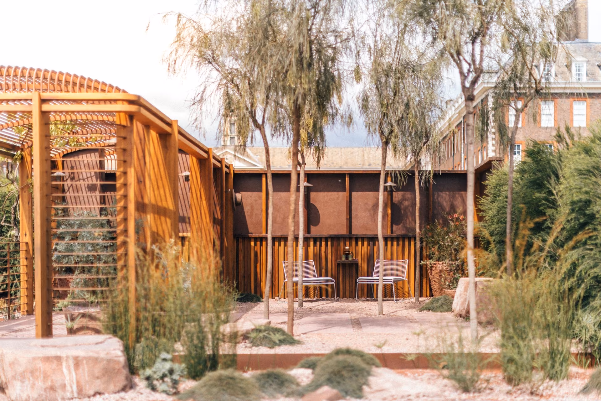 Wire chairs and a small table beneath a timber pergola in a planted courtyard garden