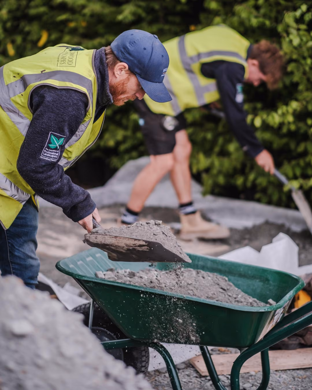 Two landscapers in hi-vis vests moving gravel with a wheelbarrow and shovels