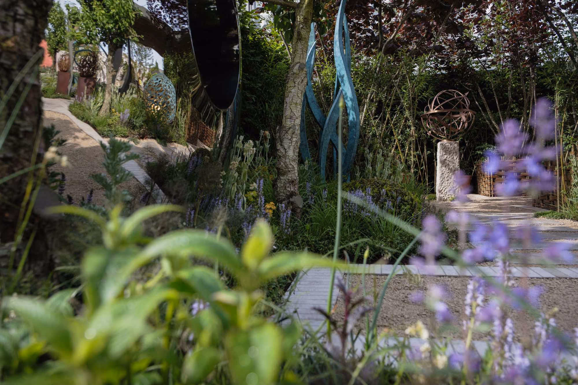 Sunlit show garden path flanked by sculptures, shrubs and purple flowering perennials