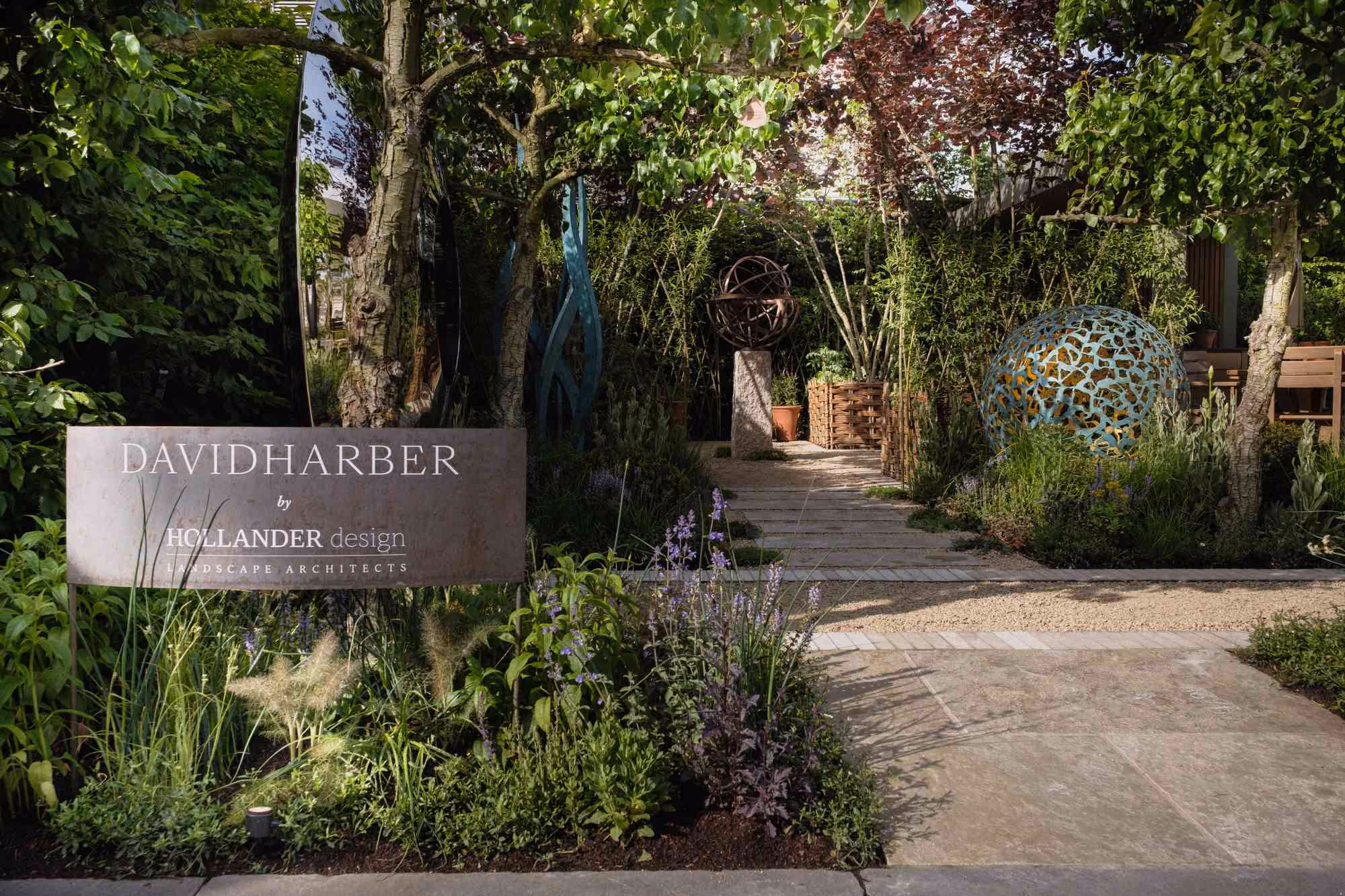 Show garden with plaques, sculptures and a stone pathway through green and purple planting