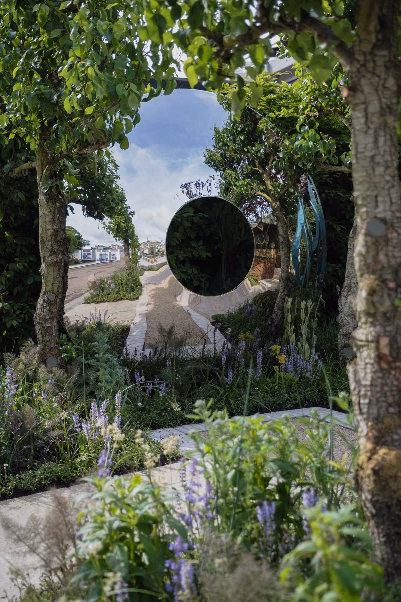 Garden path and trees reflected in a large polished circular sculpture surrounded by flowers