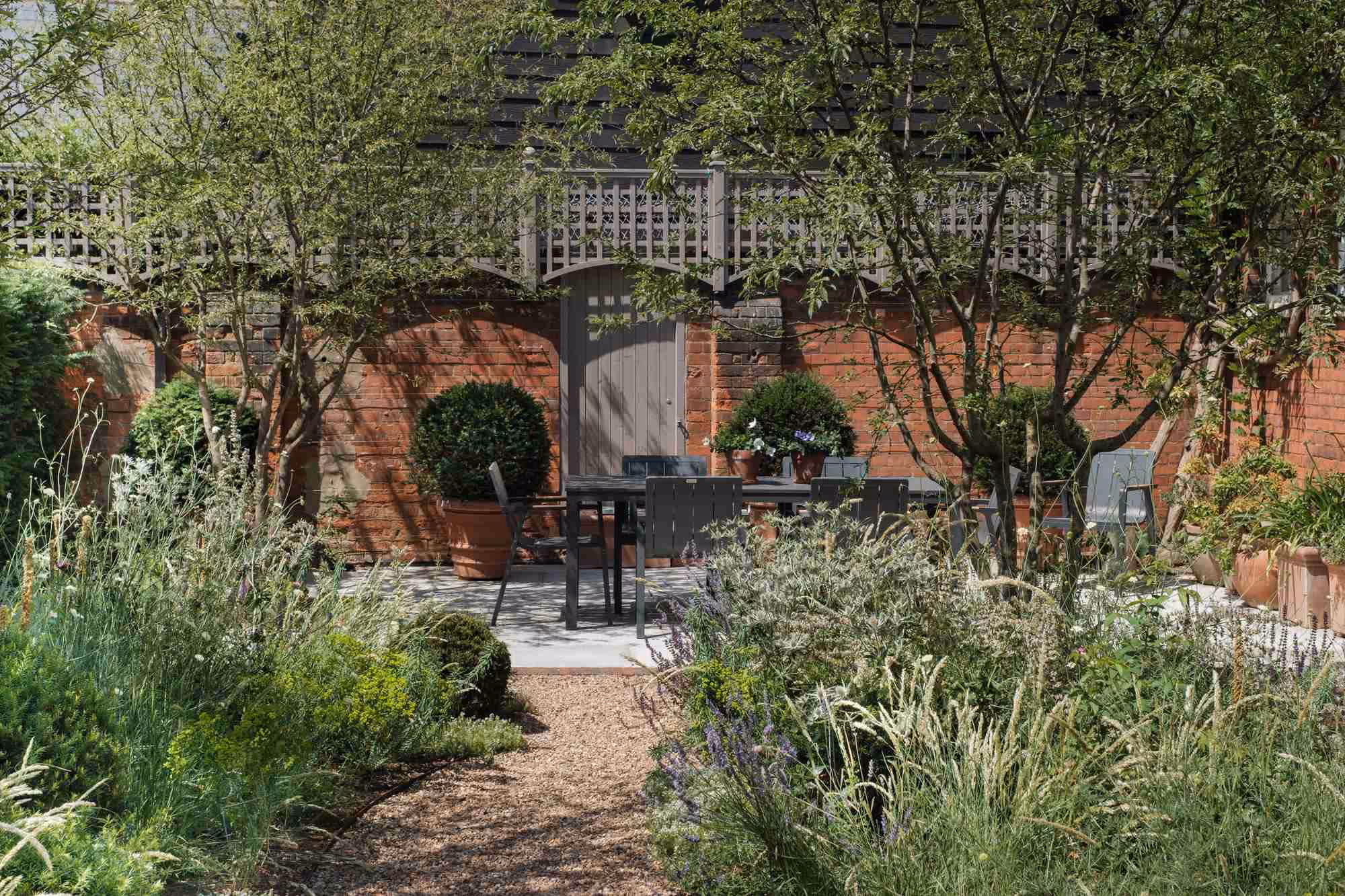 Garden path leading to a paved seating area with table, chairs and potted plants against a brick wall
