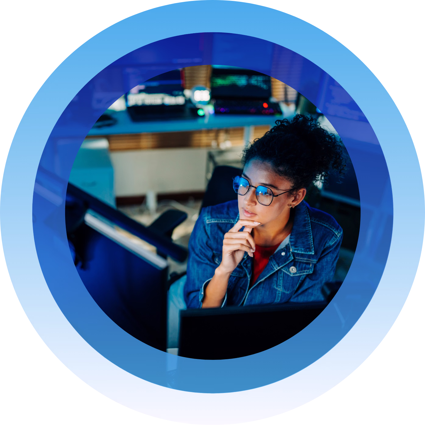 Woman with glasses and denim jacket thoughtfully looking at computer screens in a dimly lit room.