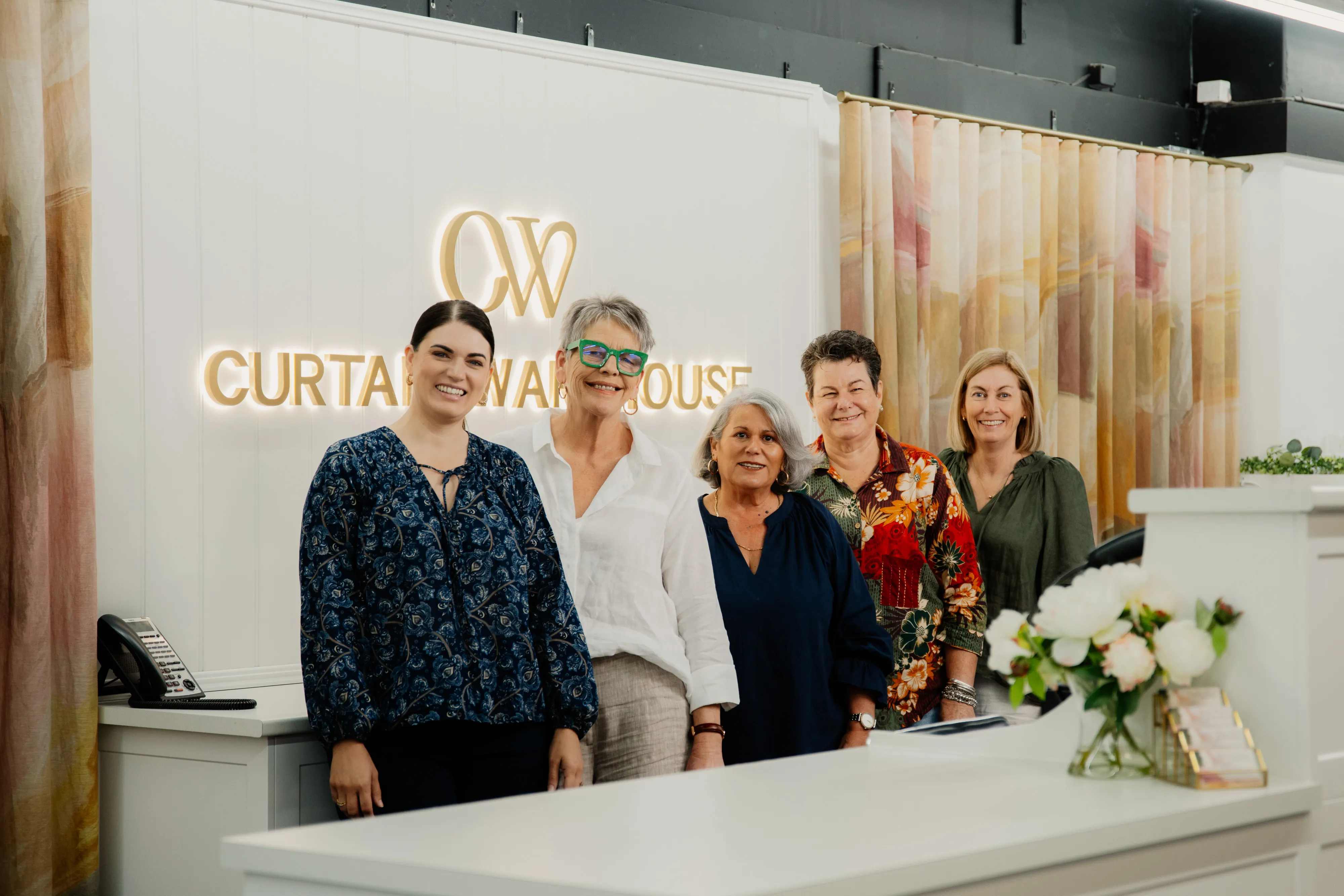 Angela Jones and her team at Curtain Warehouse & Interiors, five women standing behind a white counter with curtains and flowers in the background.