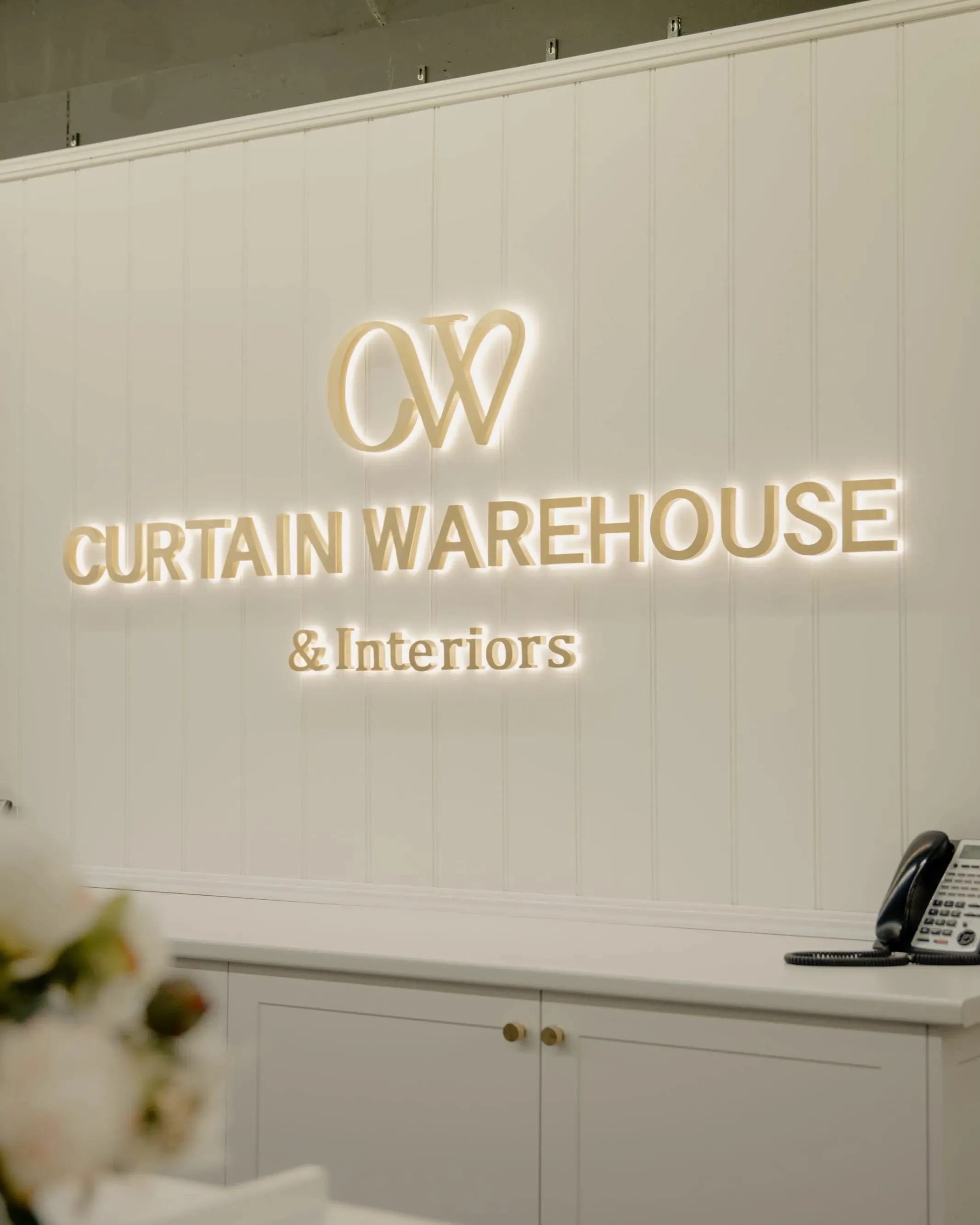 Illuminated sign reading 'CW CURTAIN WAREHOUSE & Interiors' on a white paneled wall above a counter.