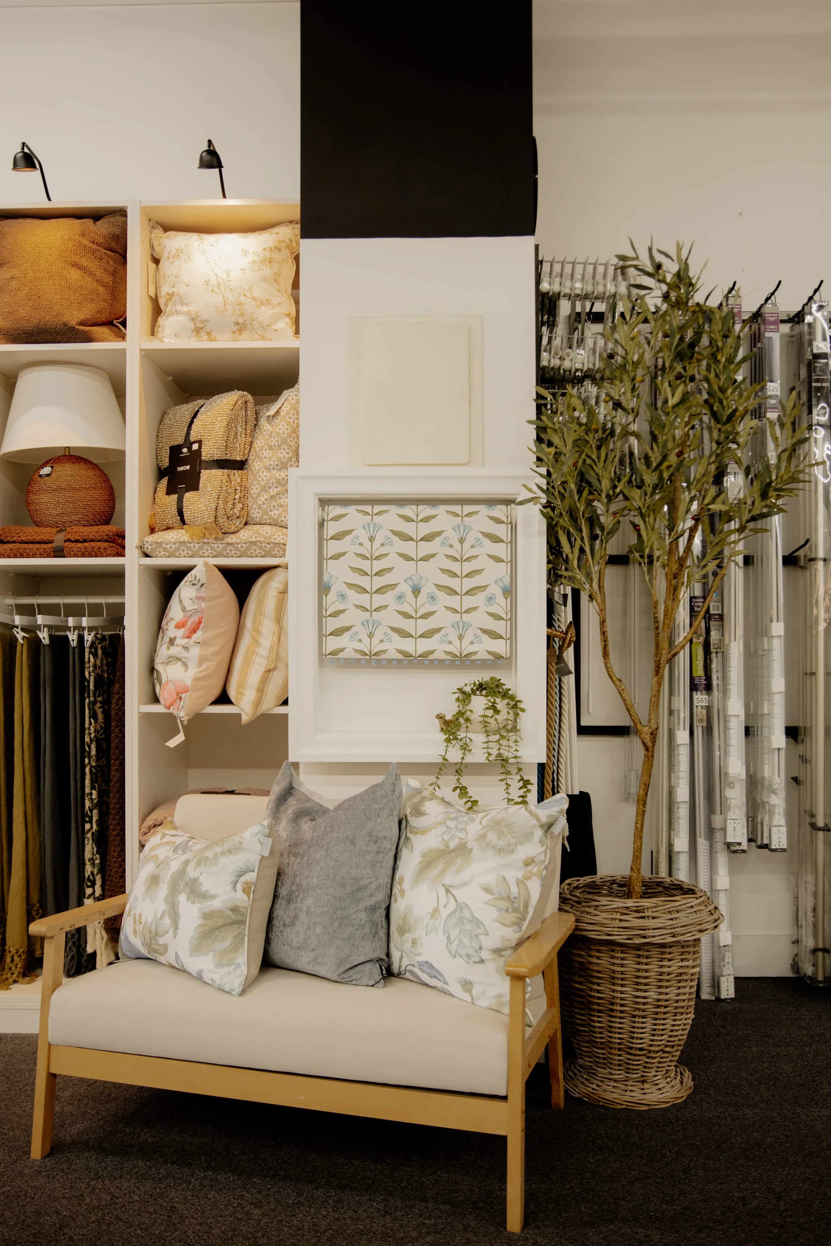 Display of cushions, fabric rolls, a wicker basket plant, and a wood-framed sofa with three pillows.