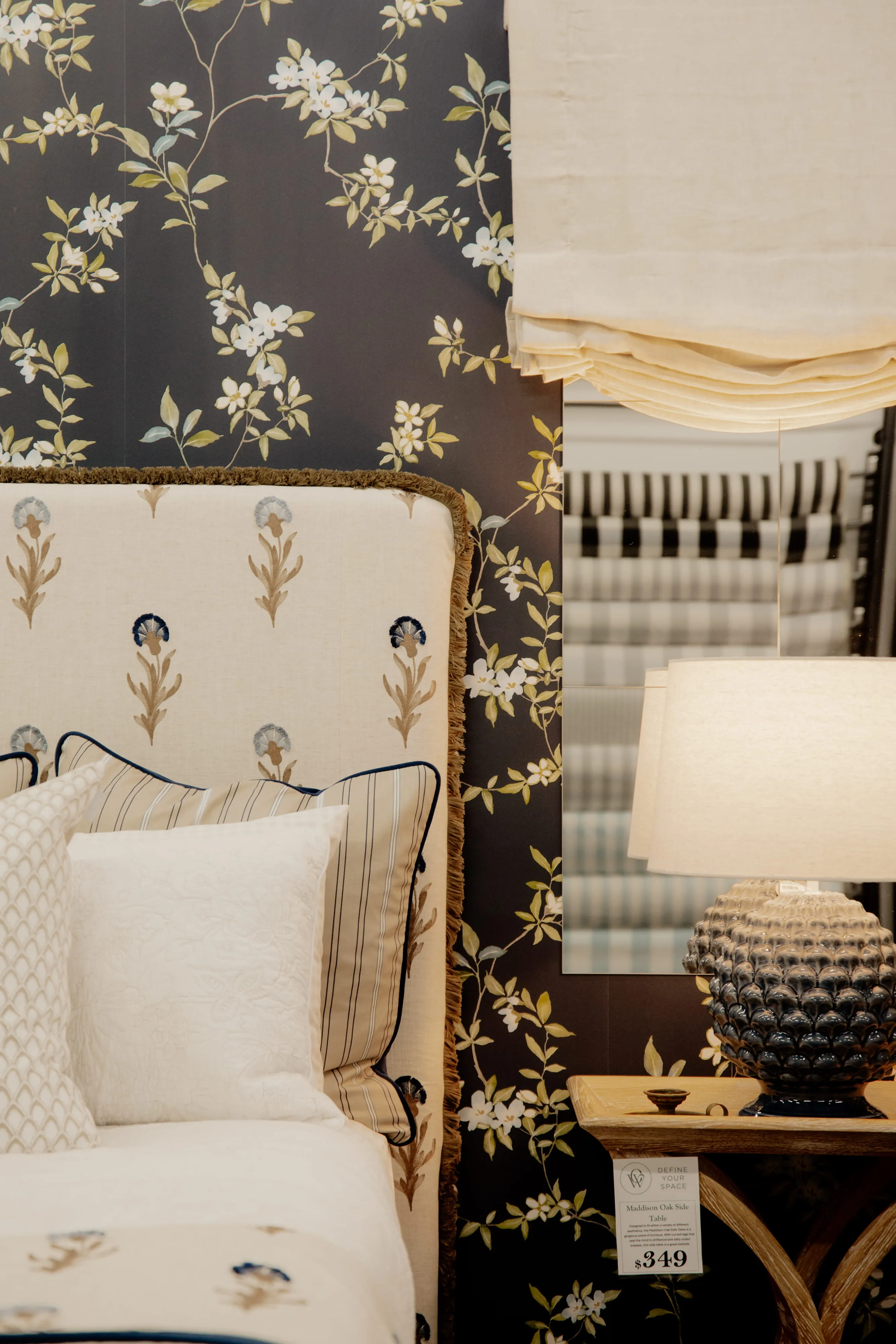 Bed with floral headboard and pillows beside a wooden table with textured lamp and floral wallpaper.