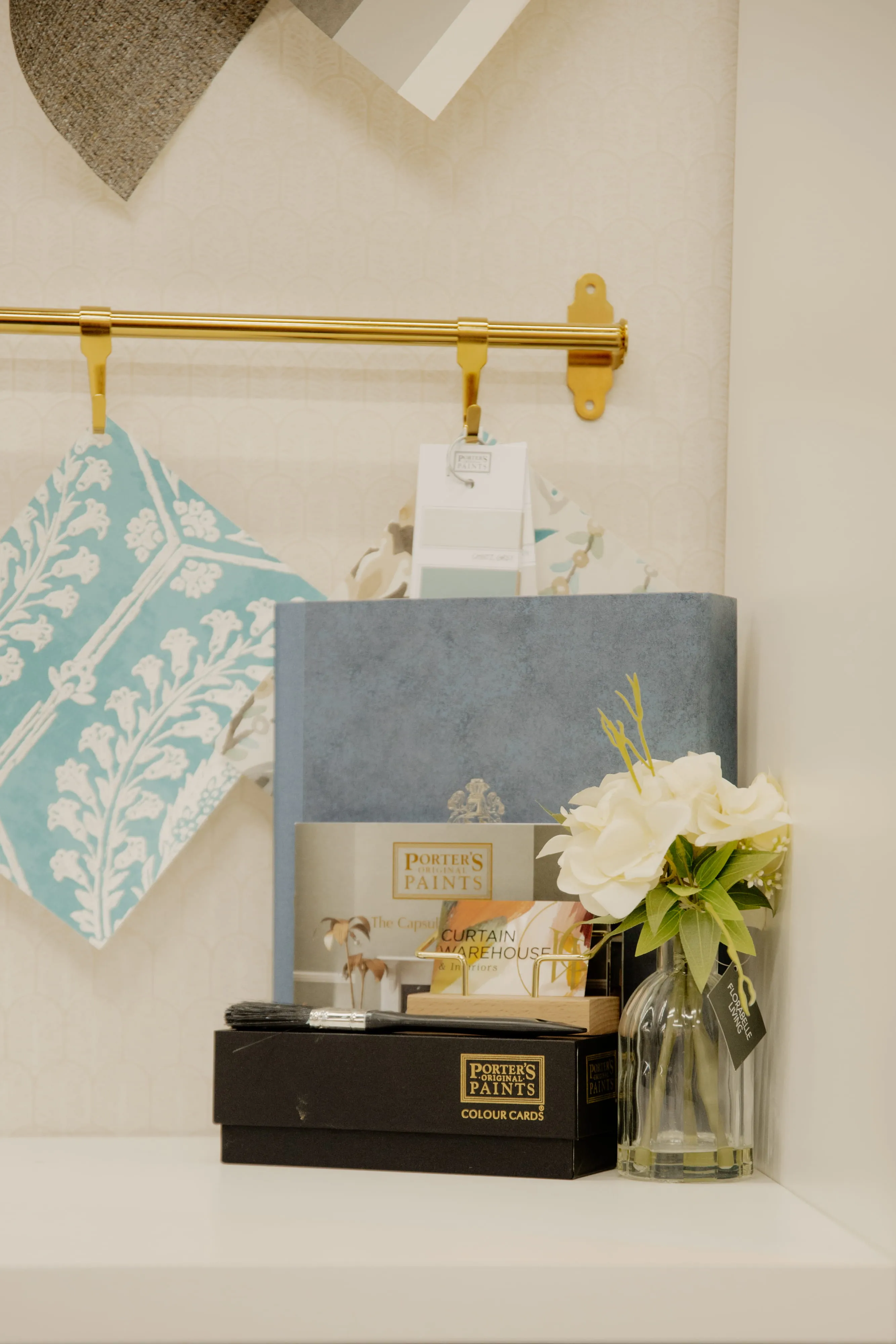 Paint color cards and brushes near hanging fabric swatches and a vase with white flowers.