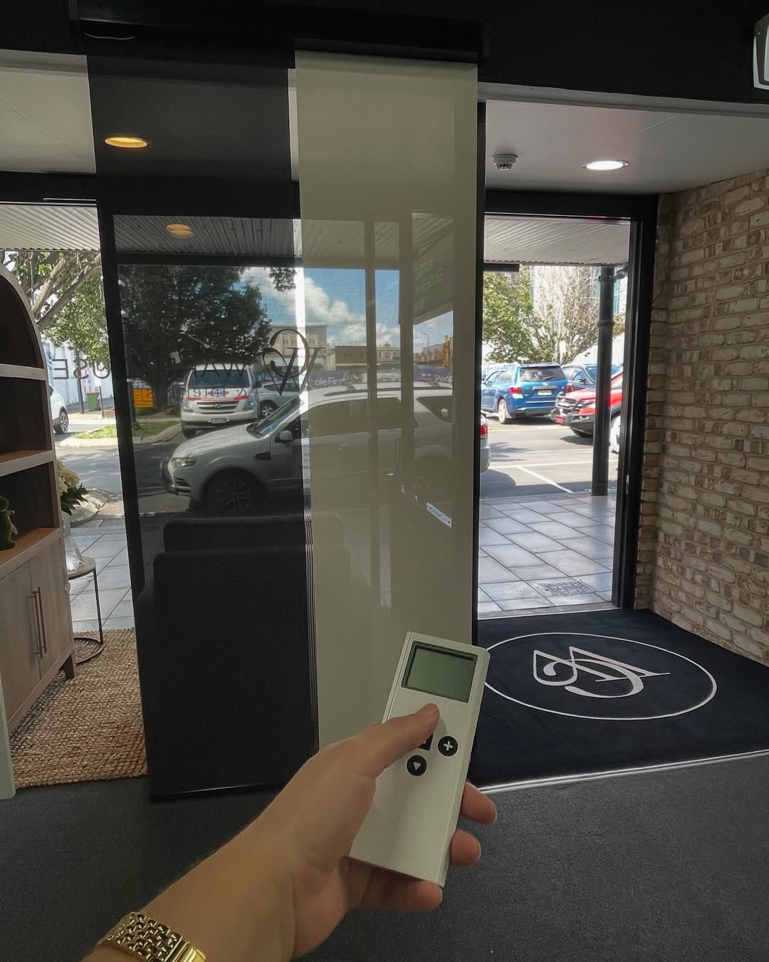Hand holding remote controlling motorised curtains in a showroom with glass doors and parked cars outside.