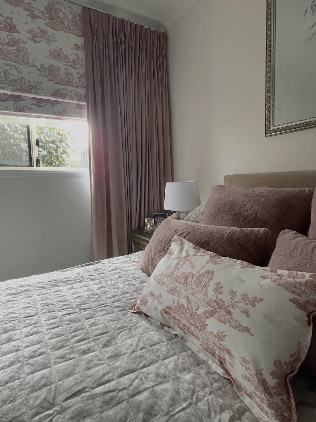 Bedroom with bed featuring pink and white pillows, pink curtains, floral roman shade, and a bedside lamp.