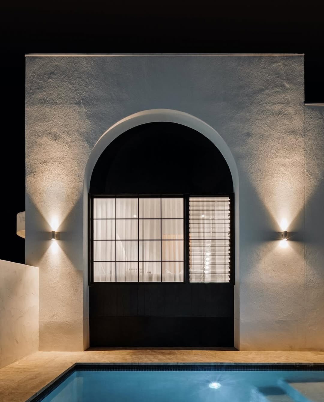 Night view of modern white building with arched window and lit swimming pool in foreground.