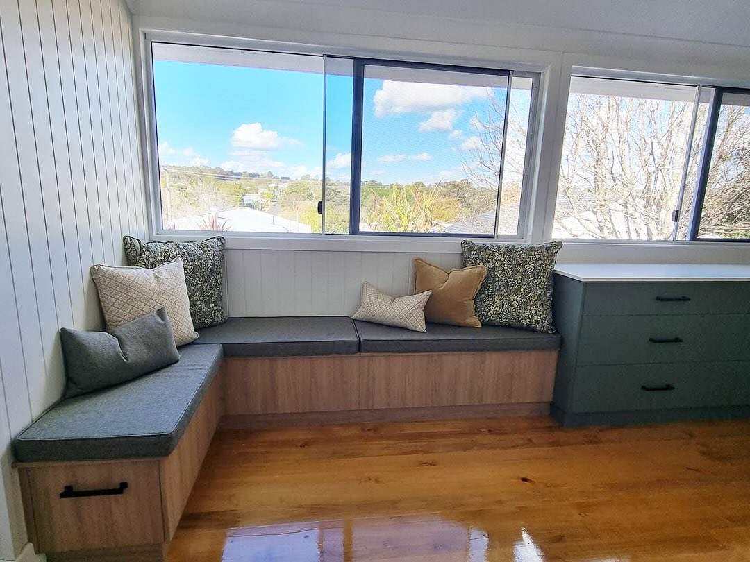 Cozy L-shaped wood bench with gray cushions and patterned pillows by large windows with landscape view.