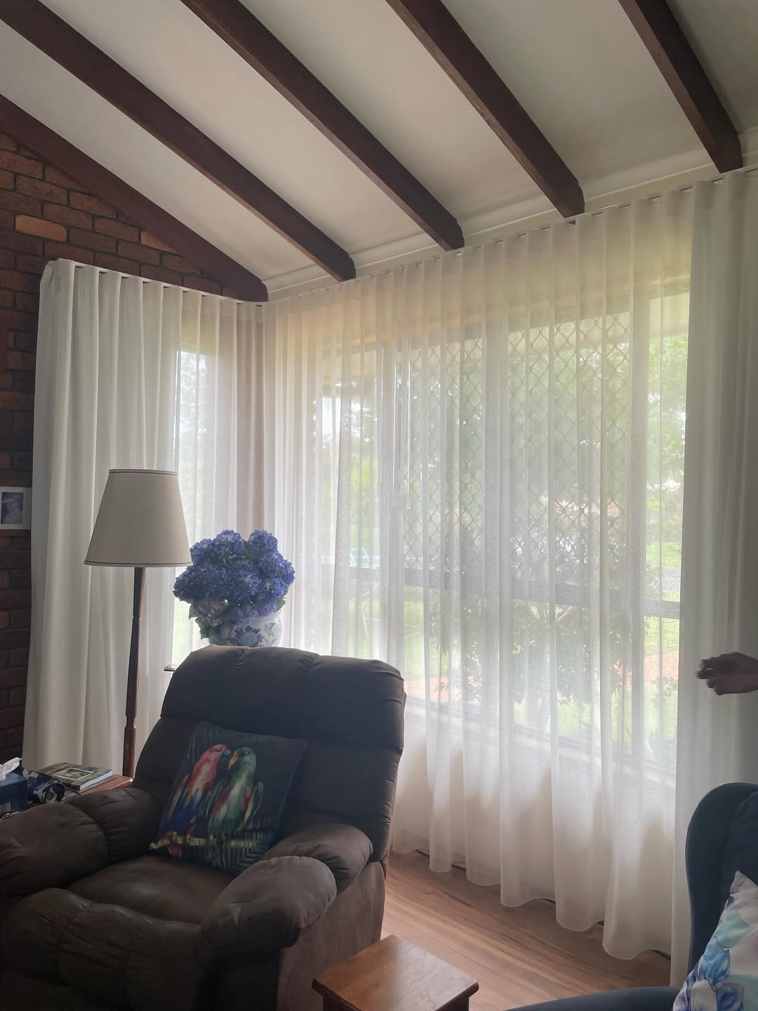 Cozy living room corner with beige sheer curtains, brown armchair, floor lamp, and blue flowers.