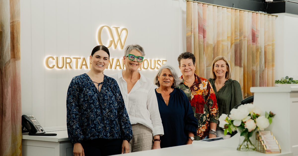 Five women standing inside a curtain store with a sign reading Curtain Warehouse behind them.