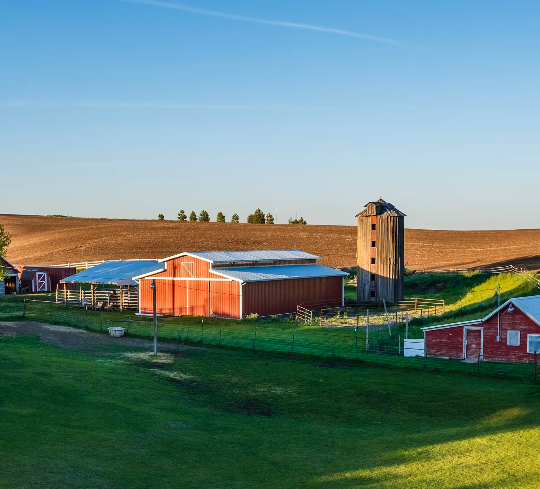 Farm with red barns, wooden silo, green grass, and a plowed field under a clear blue sky.