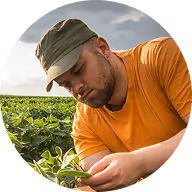 Man in orange shirt and cap inspecting green plants in a field under cloudy sky.