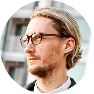 Man with light hair and glasses looking to the side in an outdoor urban setting.